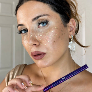 A woman with glittery makeup holds a purple cosmetic pencil in front of her face, wearing a champagne strap and gold jewelry in an indoor setting.
