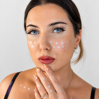 A close-up portrait of a woman with white star-shaped face glitter applied to her forehead, cheeks, and hand, wearing a black spaghetti-strap top against a light gray background.